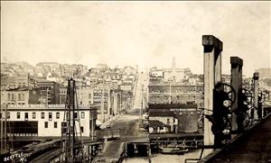 A steep hillside with rustic homes and buildings along both sides of a wide road going up the hill. In the foreground, railroad tracks built on wooden trestles curve along a shoreline. One small railcar sits on the tracks. 